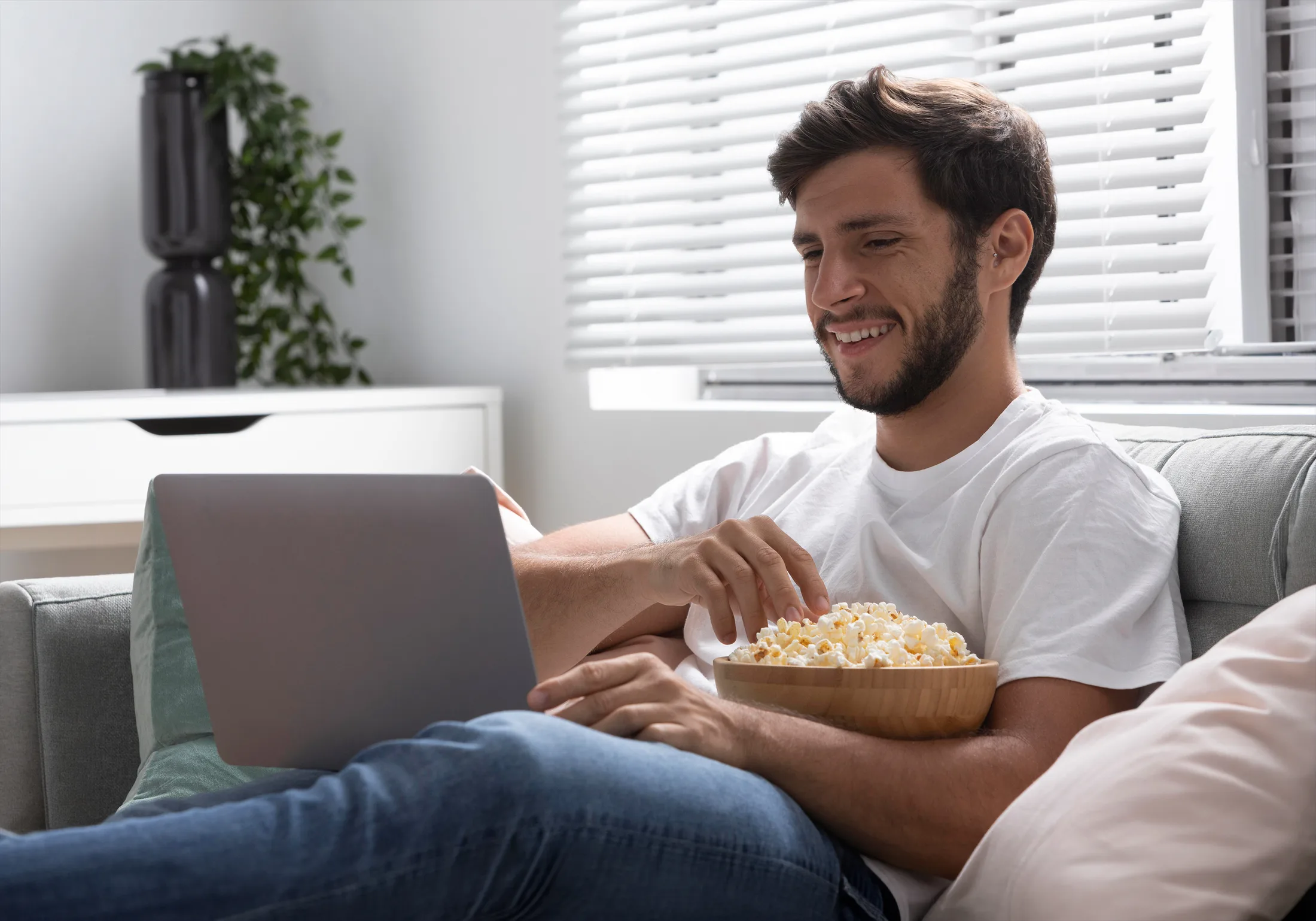 Streaming en casa: Hombre sonriendo y comiendo palomitas mientras ve una película online en su laptop.